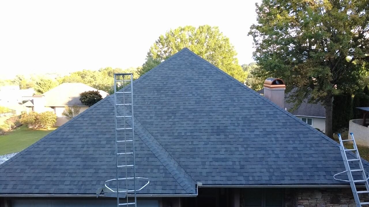 New gray architectural shingle hip roof with a gutter along the eave and two ladders set against it, trees behind